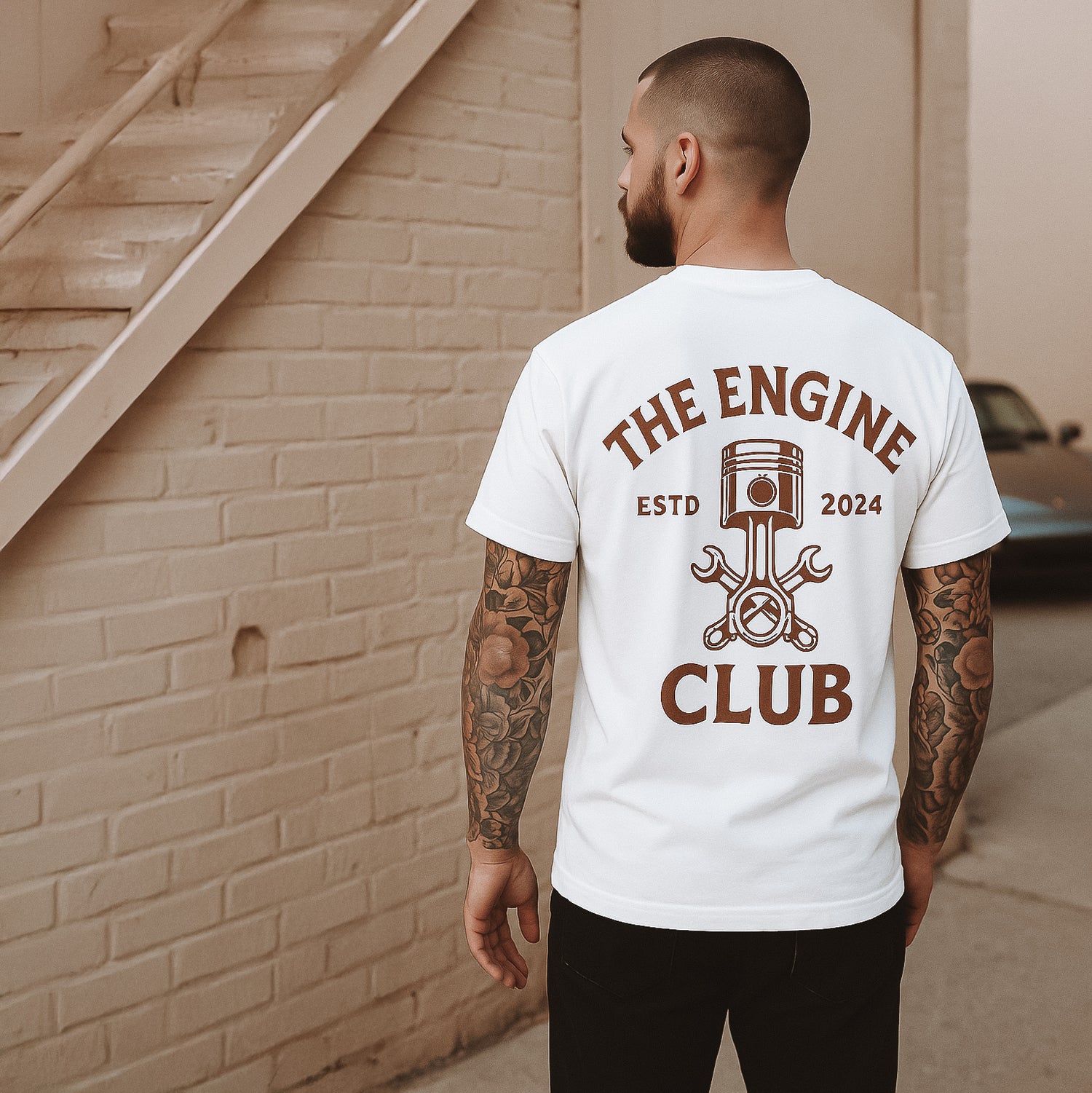 Ein tätowierter Mann steht mit dem Rücken zur Kamera und trägt ein weißes T-Shirt mit braunem Aufdruck „THE ENGINE CLUB – ESTD 2024“ sowie einem grafischen Design aus Kolben, Schraubenschlüsseln und Lenkrad; im Hintergrund eine Backsteinwand und ein geparktes Auto.